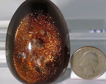 May include: A dark brown, oval-shaped resin egg with a glittery, copper-colored interior. The glitter is scattered throughout the egg, creating a swirling effect. The egg is about the size of a U.S. quarter.