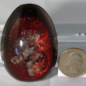 May include: A dark brown, egg-shaped resin paperweight with red glitter and white flecks embedded inside. The paperweight is about the size of a US quarter.