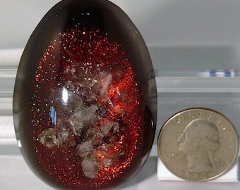 May include: A dark brown, egg-shaped resin paperweight with red glitter and white flecks embedded inside. The paperweight is about the size of a US quarter.