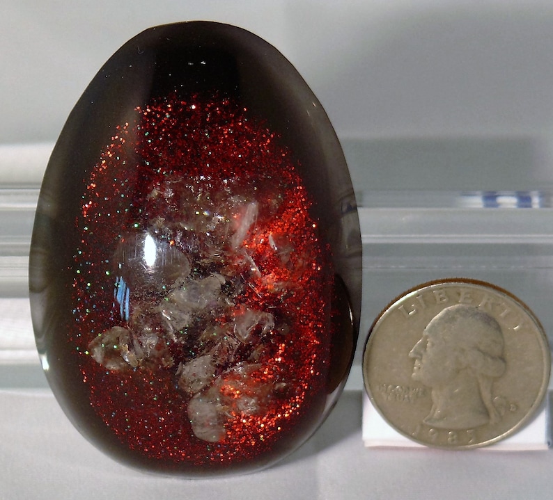 May include: A dark brown, egg-shaped resin paperweight with red glitter and white flecks embedded inside. The paperweight is about the size of a US quarter.
