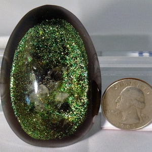 Può includere: Un fermacarte di vetro di forma ovale grigio scuro con un design di glitter verde e cristalli bianchi all'interno.