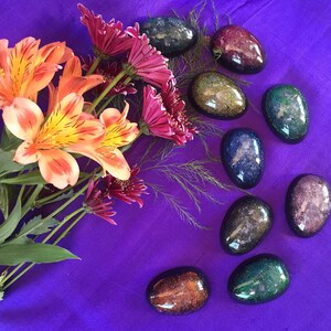 May include: A collection of eight oval-shaped resin paperweights with a galaxy-like design. The paperweights are arranged in a semi-circle on a purple surface. The paperweights are various shades of blue, green, purple, and orange.