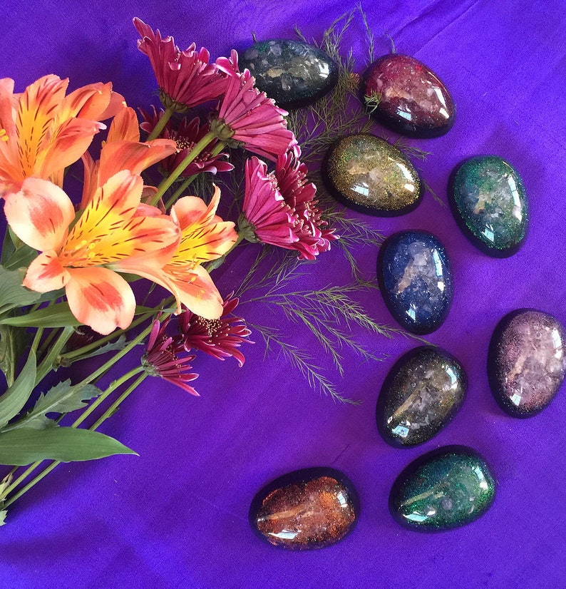 May include: A collection of eight oval-shaped resin paperweights with a galaxy-like design. The paperweights are arranged in a semi-circle on a purple surface. The paperweights are various shades of blue, green, purple, and orange.