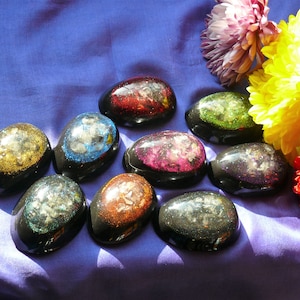 May include: A collection of ten resin paperweights, each with a different color and glitter embedded inside. The paperweights are shaped like eggs and are arranged on a purple fabric background.