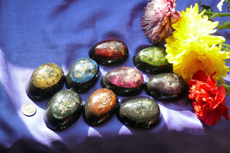 May include: A collection of ten resin paperweights, each with a different color and glitter embedded inside. The paperweights are shaped like eggs and are arranged on a purple fabric background.
