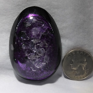 Może przedstawiać: Ozdoba w kształcie jaja z czarną obudową i fioletowym, błyszczącym wnętrzem. Przezroczyste, nieregularne kawałki są zawieszone w fioletowym rdzeniu. Obok ozdoby znajduje się moneta w celu porównania wielkości.