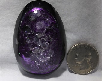 May include: An egg-shaped paperweight with a black exterior and a purple, glittery interior. Clear, irregular-shaped pieces are suspended within the purple core. A U.S. quarter is next to the paperweight for size comparison.