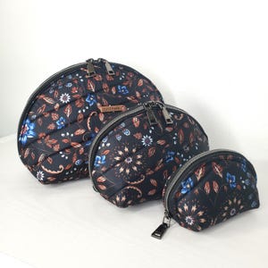 May include: Three black quilted cosmetic bags with a floral pattern in blue, white, and rust colors. The bags are different sizes, with a semi-circular shape and a zipper closure. The bags are arranged in a row.