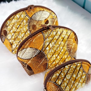 May include: Set of three oval-shaped cosmetic bags with a vibrant yellow, brown, and white patterned fabric. Each bag has a brown zipper and a matching brown trim. The bags are arranged in a descending size order.
