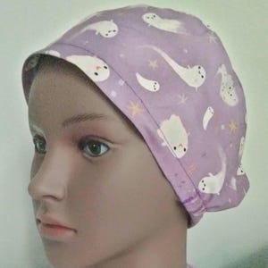 May include: A light purple scrub cap featuring a pattern of white cartoon ghosts and yellow stars. The cap is made of fabric and is designed to cover the head. The ghosts have small, black eyes and are in various poses.