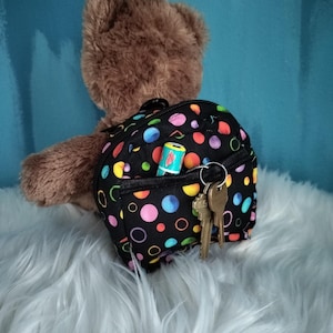 May include: A brown teddy bear wearing a small black backpack with a colorful polka dot pattern. The backpack has a front pocket with a small item and a set of keys. The bear is on a fluffy white surface against a blue background.