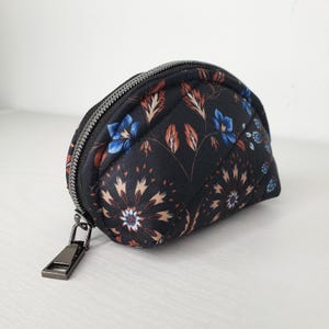 May include: A small, quilted cosmetic bag with a black background and a floral pattern in blue, brown, and cream. The bag has a silver zipper and pull. The bag is approximately 6 inches long.