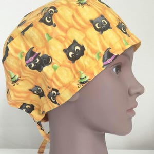 May include: A Halloween-themed surgical scrub cap featuring a repeating pattern of black cats, witch hats, and green witch hats on a yellow pumpkin background. The cap is tied at the back.
