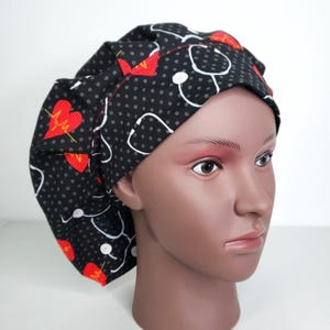 May include: A black scrub cap with a pattern of white stethoscopes, red hearts, and gray polka dots. The cap is designed to cover the hair and is likely used in a medical setting. The cap is on a mannequin head.