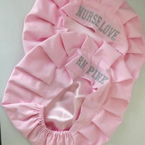 May include: Two pink satin-lined bouffant scrub caps. The larger cap has "NURSE LOVE" embroidered in silver, and the smaller cap has "RN PINK" embroidered in silver. The caps are designed for healthcare professionals.