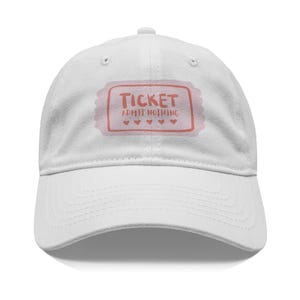 May include: White baseball cap with a curved brim. The cap features a pink ticket design with the words "TICKET ADMIT NOTHING" and four small hearts. The cap is made of a soft fabric.