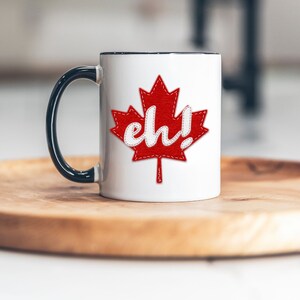 May include: White ceramic mug with a black rim and handle. The mug features a red felt maple leaf with white stitching and the word "eh!" in white felt.