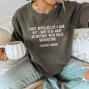 May include: Dark gray t-shirt with the quote "I HAVE NEVER KILLED A MAN, BUT I HAVE READ MANY AN OBITUARY WITH GREAT SATISFACTION." attributed to Clarence Darrow. The shirt is worn with light blue jeans. A white pumpkin-shaped mug is held in the hand.