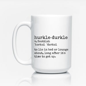 May include: White ceramic mug with a black and white definition of the word "hurkle-durkle" printed on it. The definition is a Scottish word meaning to lie in bed or lounge about long after it's time to get up.