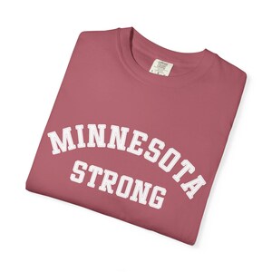 May include: A folded, dusty rose-colored t-shirt with the words "MINNESOTA STRONG" in white, arched lettering. The shirt has a crew neck and a small tag on the upper back.