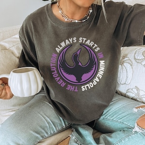 May include: Dark grey t-shirt with a circular graphic in purple and black, featuring a bird design and the text "ALWAYS STARTS IN MINNEAPOLIS THE REVOLUTION". The person is holding a white pumpkin-shaped mug.