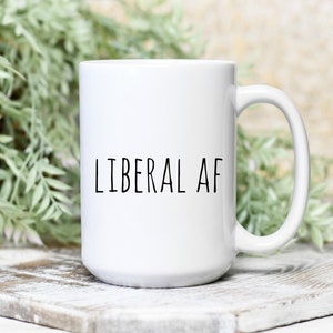 May include: White ceramic mug with the text "LIBERAL AF" in black, centered on the front. The mug has a large handle and is set against a blurred background of greenery and a white wooden surface.