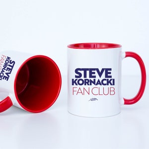 May include: Two white ceramic mugs with red rims. The mug on the right has the text "STEVE KORNACKI FAN CLUB" in blue lettering with a small sprig of wheat underneath.