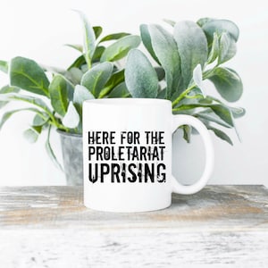 May include: White ceramic mug with black text that reads "Here for the Proletariat Uprising". The mug is sitting on a wooden surface with a green plant in the background.