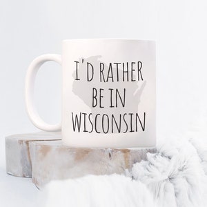 May include: A white ceramic mug featuring a light gray silhouette of the state of Wisconsin in the background. The text "I'D RATHER BE IN WISCONSIN" is printed in a simple, black, sans-serif font across the center of the mug.
