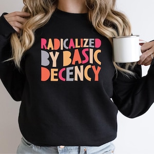 Op de afbeelding: Zwart sweatshirt met de tekst "RADICALIZED BY BASIC DECENCY" in kleurrijke blokletters. Het sweatshirt is effen zwart van kleur. Een witte mok wordt in de hand gehouden.