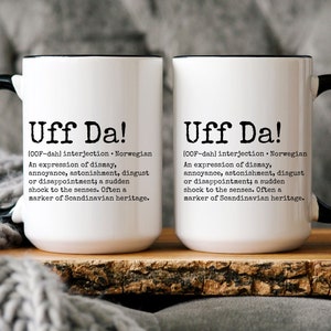 May include: Two white ceramic mugs with black handles and black rims. Both mugs feature the text "Uff Da!" with a definition of the word in black text. The definition is "[OOF-dah] interjection Norwegian An expression of dismay, annoyance, astonishment, disgust or disappointment; a sudden shock to the senses. Often a marker of Scandinavian heritage."