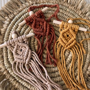 Może przedstawiać: Trzy ręcznie robione lalki z makramy w odcieniach beżu, rdzy i musztardowej żółci. Każda lalka ma drewnianą głowę i ramiona, z wiązanym korpusem i frędzlami. Lalki ułożone są na okrągłym, plecionym podkładzie.