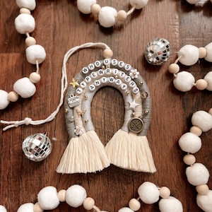 Może przedstawiać: Wisząca ozdoba ścienna z makramu w kolorze szarym i białym z napisem "Who Uses Typewriters Anyways" czarnymi literami. Ozdoba jest ozdobiona koralikami, gwiazdami i zawieszką w kształcie maszyny do pisania.