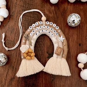 Puede incluir: Un tapiz de macramé arcoíris beige y marrón con las palabras "All's Fair in Love and Poetry" escritas en cuentas blancas. El arcoíris tiene un amuleto de llave, un amuleto de estrella y un amuleto de flor.