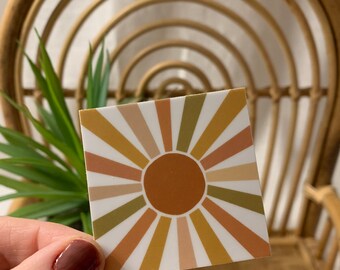 May include: A square sticker with a sunburst design in shades of brown, beige, and green. The sunburst has a large, solid brown circle in the center.