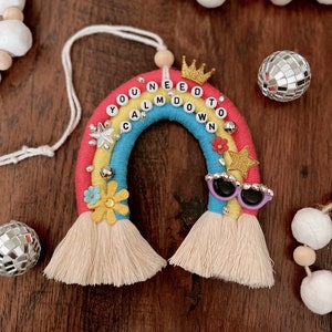 Op de afbeelding: Een regenboogvormig ornament met de tekst "YOU NEED TO CALM DOWN" in letterkralen. De regenboog heeft rode, gele en blauwe secties, versierd met een gouden kroon, ster, zonnebril en kwasten. Decoratieve kralen en discoballen zijn ook aanwezig.