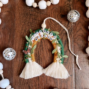 Puede incluir: Una pared colgante de macramé verde, amarilla y blanca con las palabras "Life Was A Willow" escritas en cuentas blancas. La pared colgante tiene un amuleto de mariposa y acentos de estrella dorada.