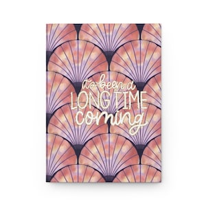 May include: A hardcover journal with a repeating fan-shaped pattern in shades of pink, peach, and purple. The text "it's been a LONG TIME coming" is in a cursive font across the front cover.