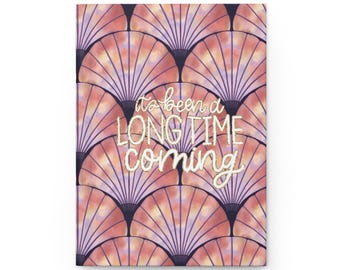 May include: A hardcover journal with a repeating fan-shaped pattern in shades of pink, peach, and purple. The text "it's been a LONG TIME coming" is in a cursive font across the front cover.