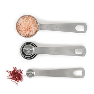 May include: Three stainless steel measuring spoons with handles. The largest spoon is filled with pink Himalayan salt. The middle spoon is labeled "1/4 tsp/1.25ml". The smallest spoon is labeled "1/8 tsp/0.63ml".