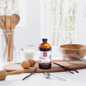 May include: A brown glass bottle of pure single fold vanilla extract from the SLO FOOD GROUP. The bottle is sitting on a wooden cutting board with vanilla beans, two brown eggs, a measuring cup, a wooden rolling pin, and a whisk.