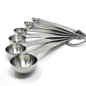May include: A set of six stainless steel measuring spoons with a hanging loop. The spoons are labeled with measurements in both teaspoons and tablespoons.