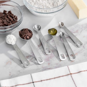 May include: A set of six stainless steel measuring spoons with different sizes, including 1/4 tsp, 1/2 tsp, 1 tsp, 1/8 tsp, 1/4 tbsp, and 1/2 tbsp. The spoons are laid out on a white marble surface.