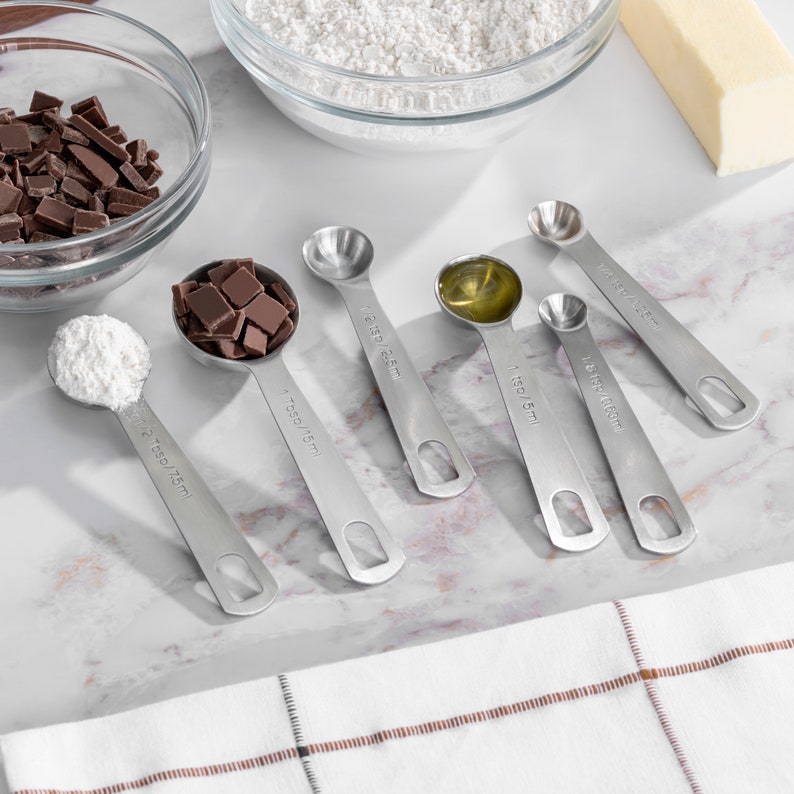 May include: A set of six stainless steel measuring spoons with different sizes, including 1/4 tsp, 1/2 tsp, 1 tsp, 1/8 tsp, 1/4 tbsp, and 1/2 tbsp. The spoons are laid out on a white marble surface.
