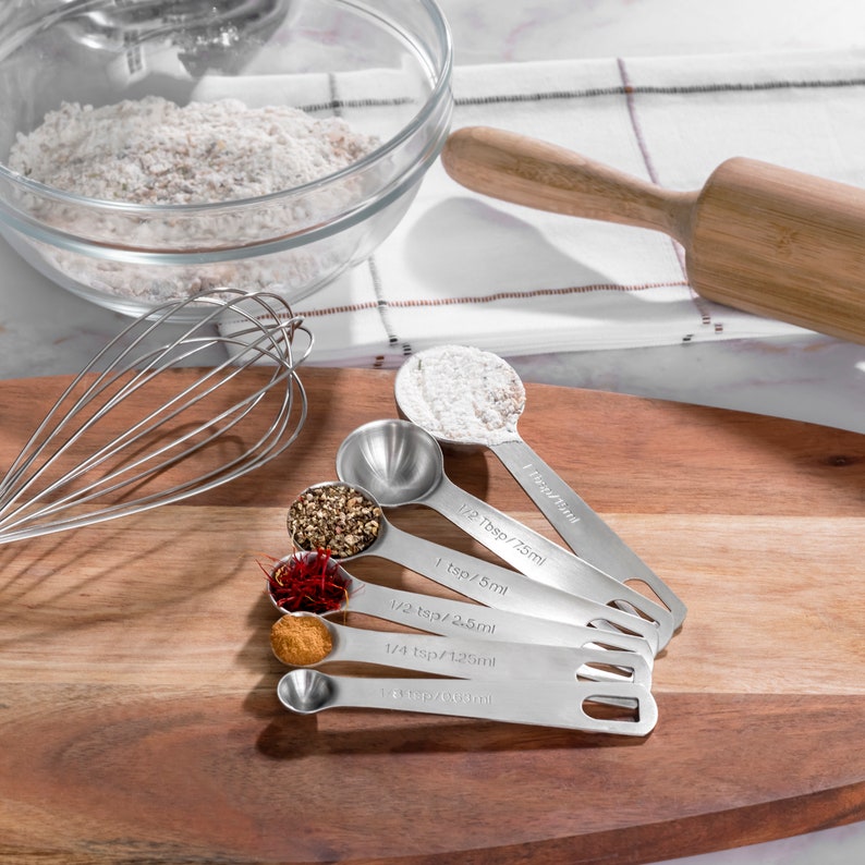 May include: A set of six stainless steel measuring spoons on a wooden cutting board. The spoons are arranged in order of size, with the largest spoon at the top and the smallest spoon at the bottom. The spoons are filled with various spices, including cinnamon, saffron, peppercorns, and powdered sugar.