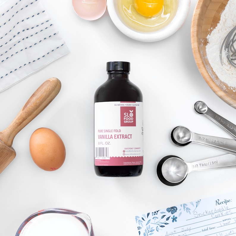 May include: A dark brown glass bottle of pure single fold vanilla extract, labeled "SLO FOOD GROUP" and "VANILLA EXTRACT", with a white background and baking supplies, including measuring spoons, a rolling pin, eggs, and a bowl of flour.