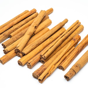 May include: A pile of brown cinnamon sticks on a white background. The sticks are arranged in a random pattern, with some overlapping.