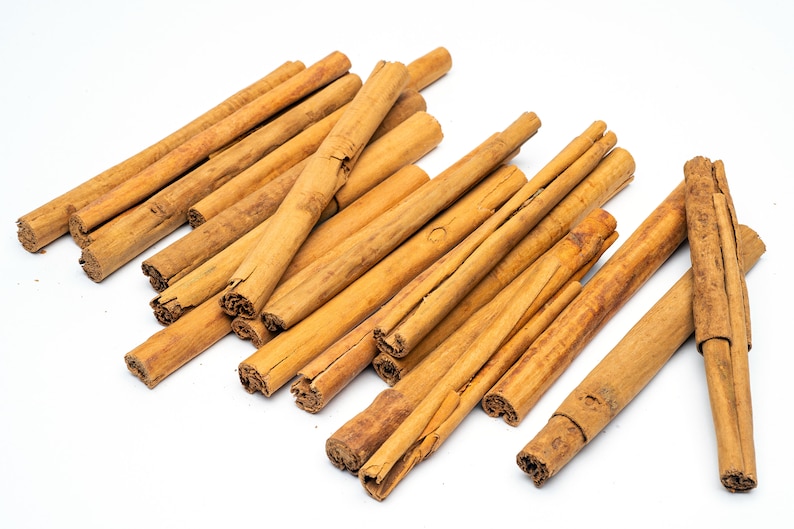 May include: A pile of brown cinnamon sticks on a white background. The sticks are arranged in a random pattern, with some overlapping.