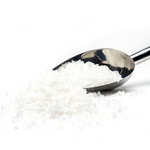 May include: A silver scoop filled with white coarse salt, spilling out onto a white surface.