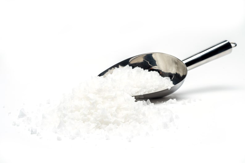 May include: A silver scoop filled with white coarse salt, spilling out onto a white surface.